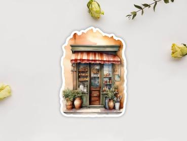 Sticker mit der Fassade eines kleinen Ladens im mediterranen Stil, dekoriert mit großen Pflanzen in Terrakottatöpfen vor dem Eingang. Eine rot-weiß gestreifte Markise bedeckt die Eingangstür, durch die Regale mit Keramik und anderen Gegenständen sichtbar
